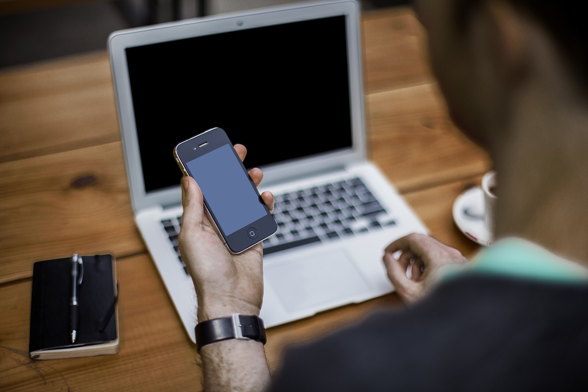 Pessoa segurando um celular diante de um notebook em uma mesa de trabalho